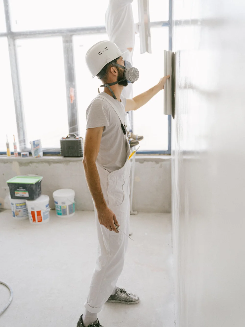 Peintre en tenue blanche appliquant la peinture sur un mur près d'une fenêtre.